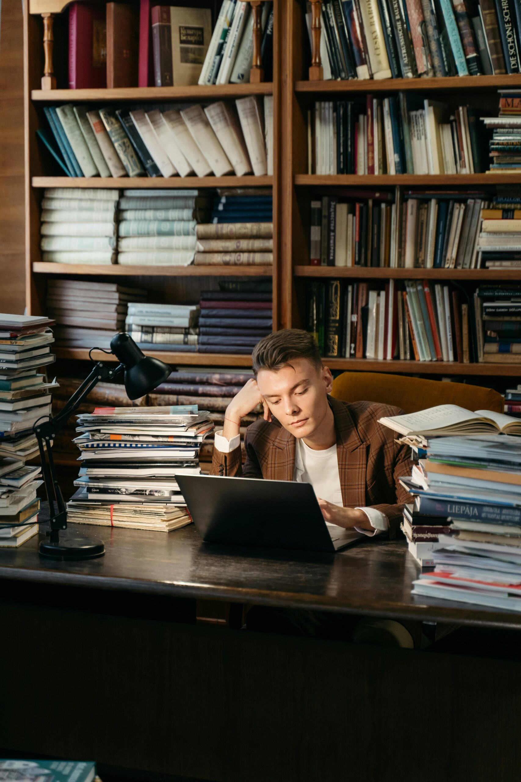A man in a suit jacket studies in a library, surrounded by books and using a laptop.